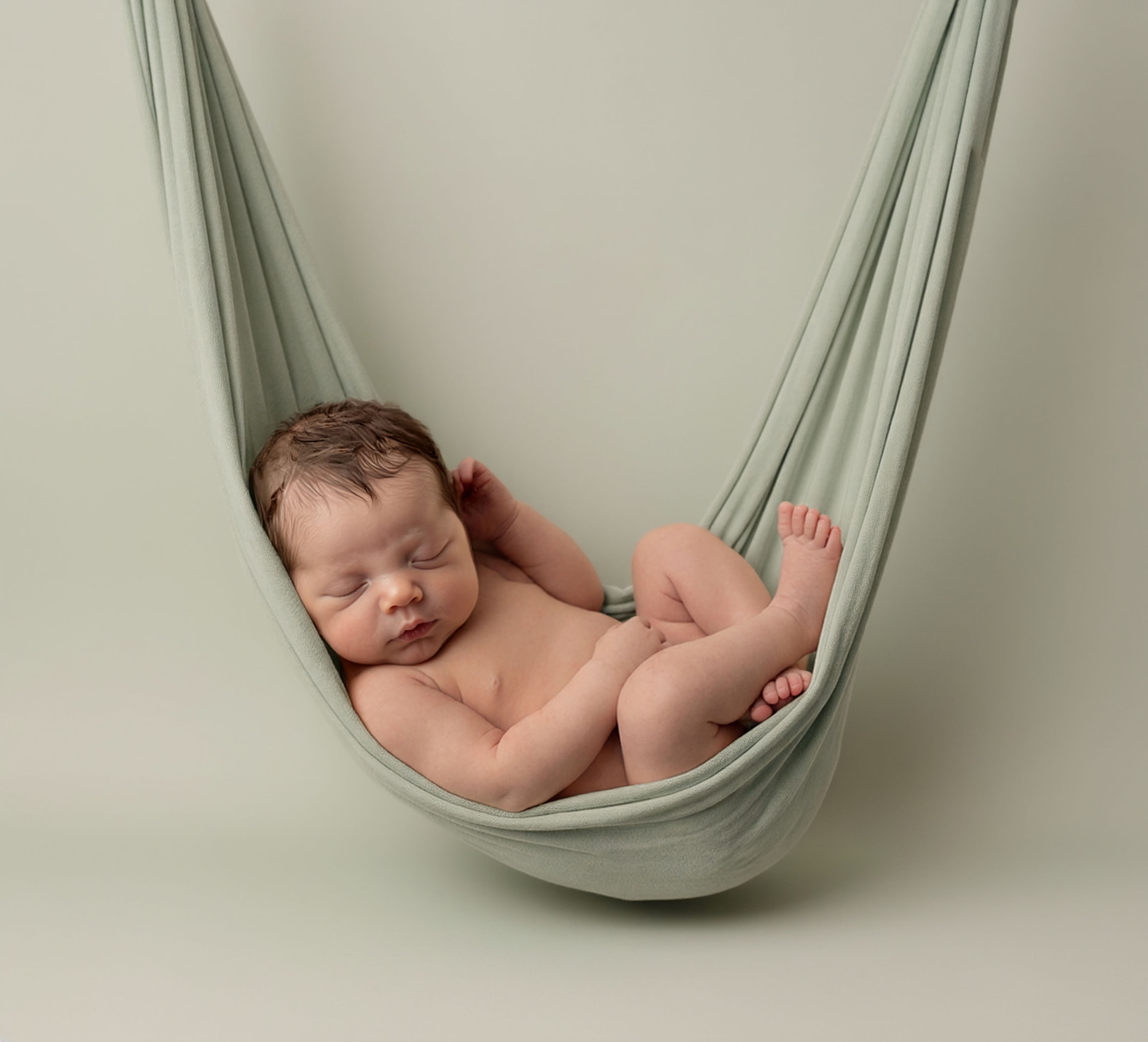 Sleeping newborn with dark wispy hair curled inside a soft sage green fabric hammock against a matching sage backdrop